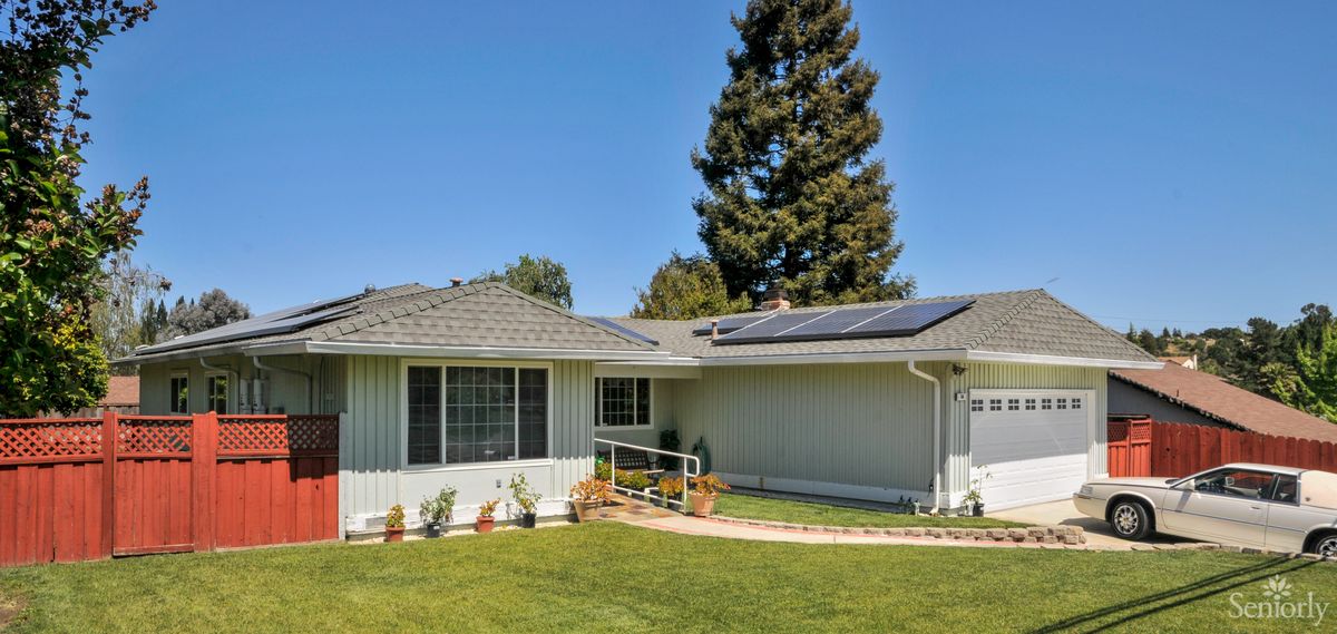 A single-story house with solar panels on its roof, surrounded by a well-maintained lawn and trees on a sunny day.
