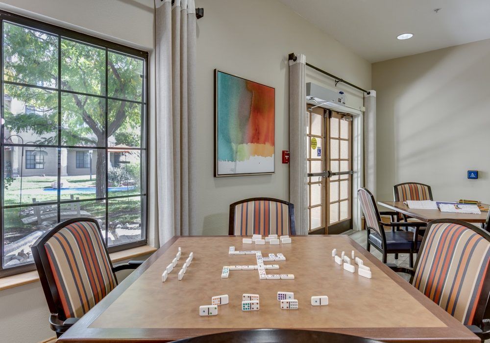 A cozy indoor room with a table set up for a game of dominoes, surrounded by chairs and natural light streaming through the window.
