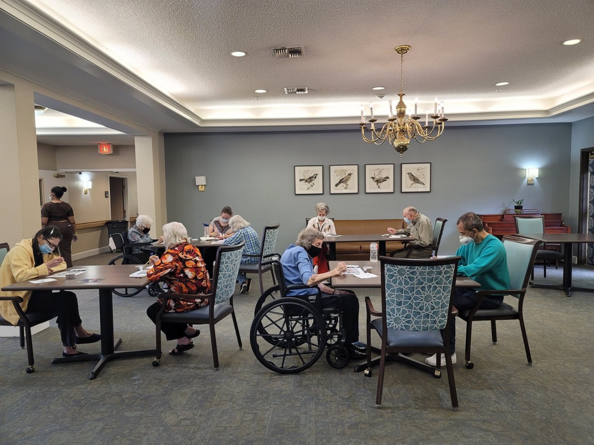 A group of seniors gathered in a communal space, some sitting in wheelchairs and others at tables.
