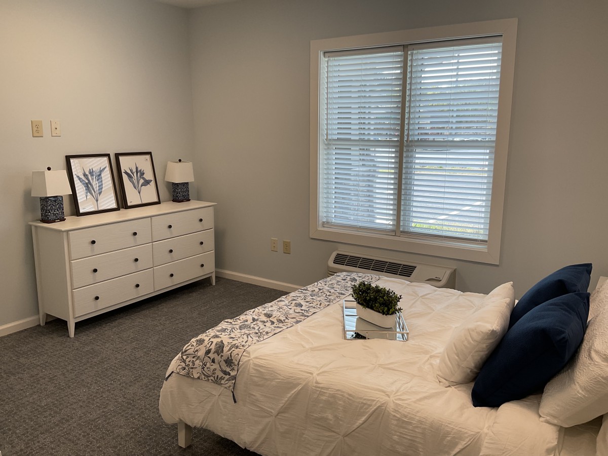 A minimalist bedroom with a neatly made bed, dresser, and a window with blinds.