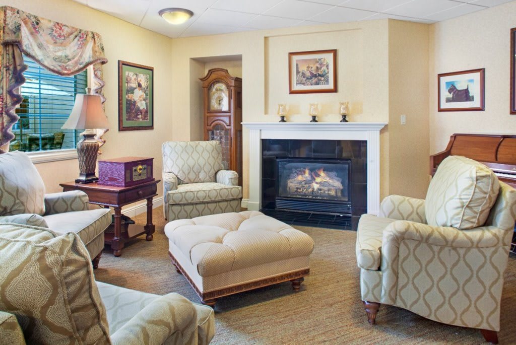 A cozy living room with a fireplace, armchairs, and an ottoman, featuring traditional decor and framed artwork.