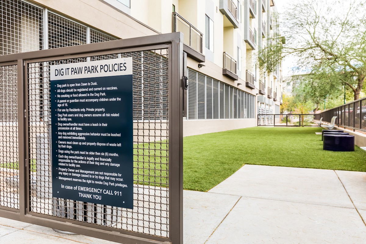An outdoor dog park area adjacent to an apartment building, featuring a set of rules on a sign attached to the fence.