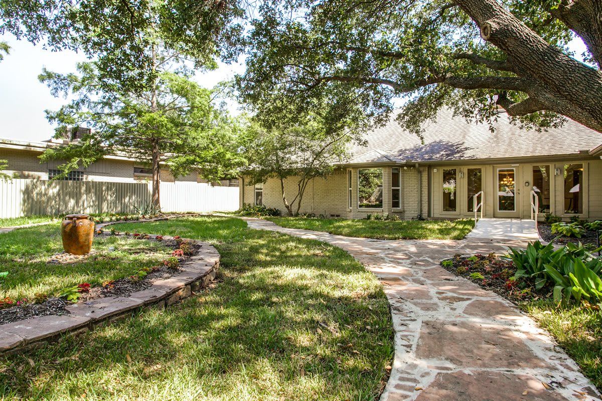 A charming house with a stone pathway winding through a well-maintained garden and lawn, shaded by large trees.