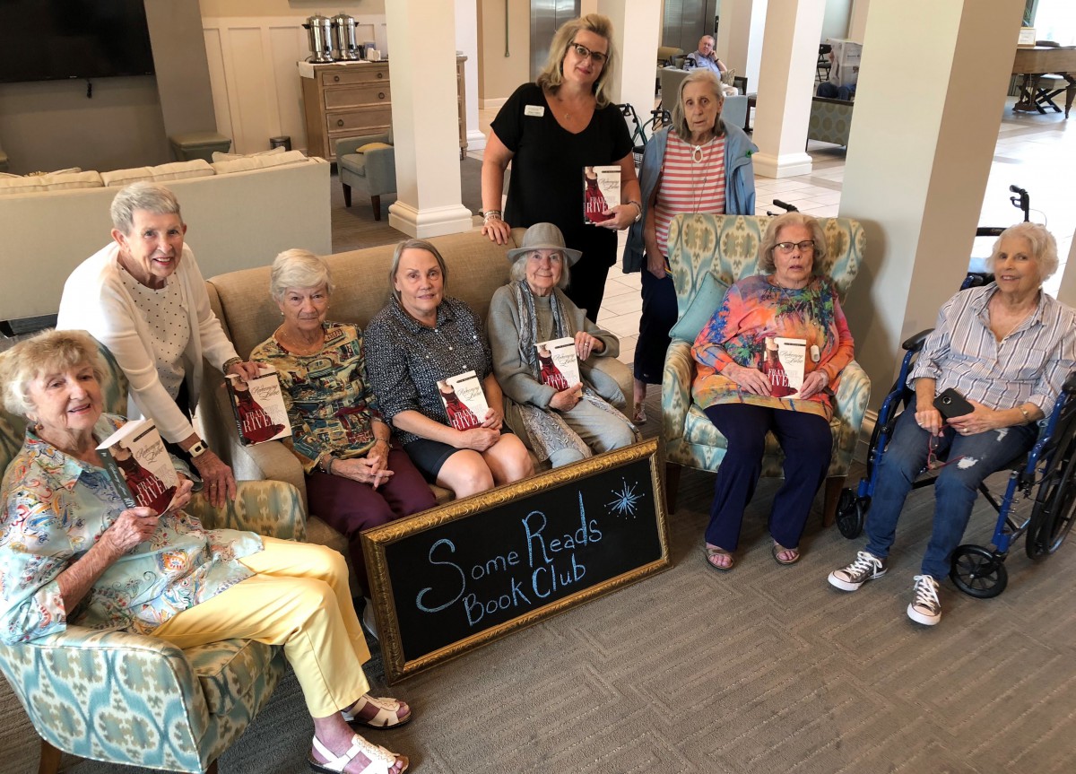 A group of elderly women holding books, seated together indoors with a sign in front of them.
