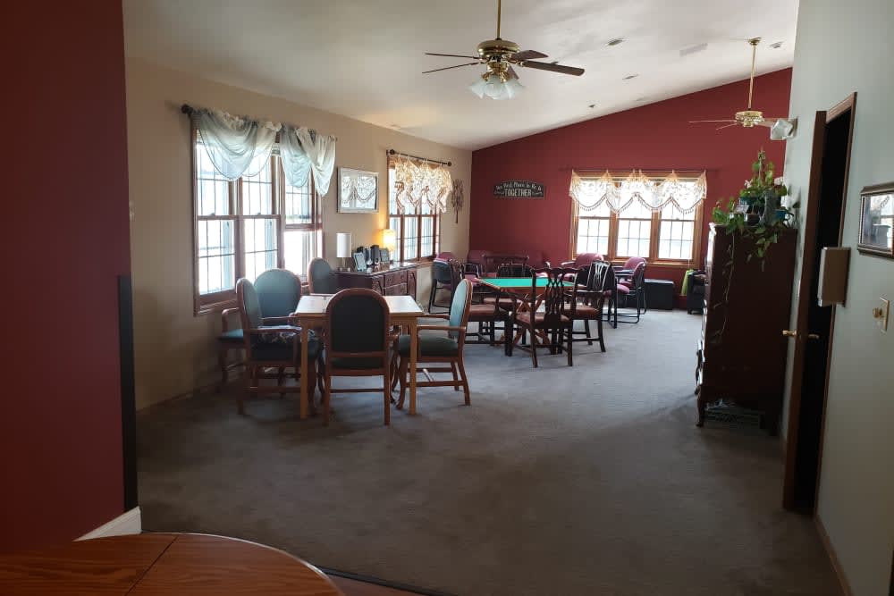 A spacious living room with tables and chairs, featuring large windows and a ceiling fan.