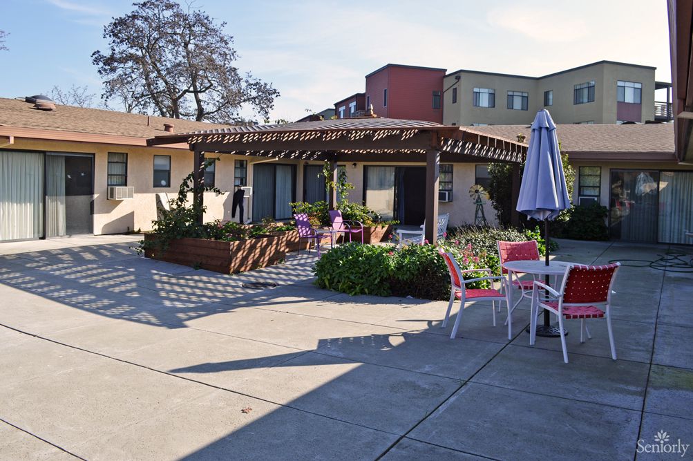 A sunny outdoor patio with tables, chairs, and a pergola surrounded by plants and buildings.