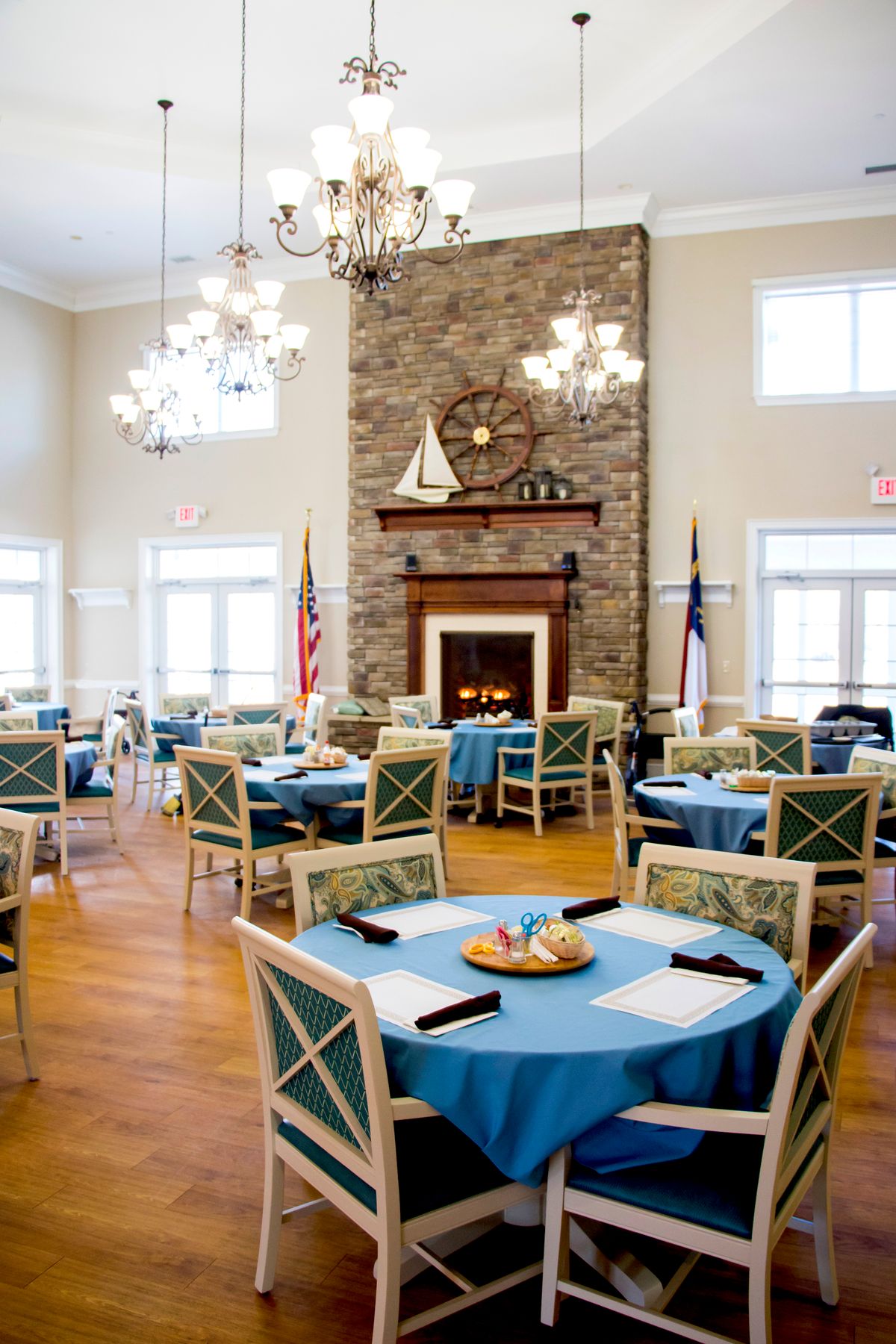 A cozy dining area with a stone fireplace, chandeliers, and neatly arranged tables covered with blue tablecloths.