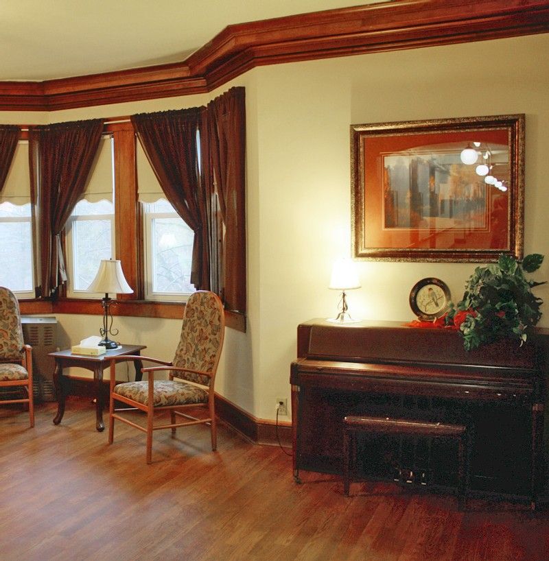 A cozy living room featuring a piano, two chairs by a window with curtains, and artwork on the wall.
