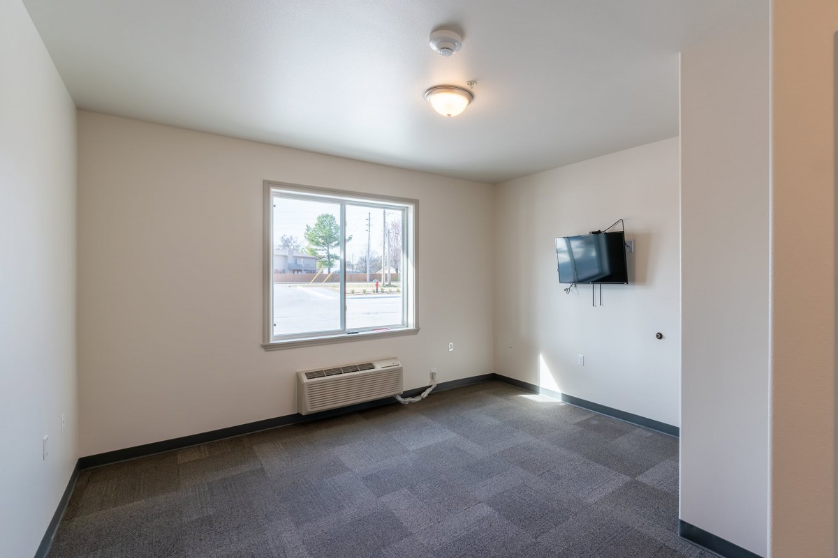 An empty room with a window, a wall-mounted TV, and carpeted flooring.