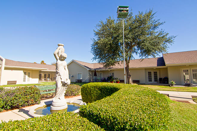 A serene garden scene featuring a classical statue, neatly trimmed bushes, and a single-story house in the background under a clear blue sky.