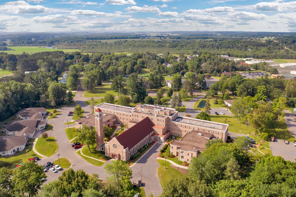 Aerial view of a church surrounded by lush greenery and a parking lot, set under a partly cloudy sky.