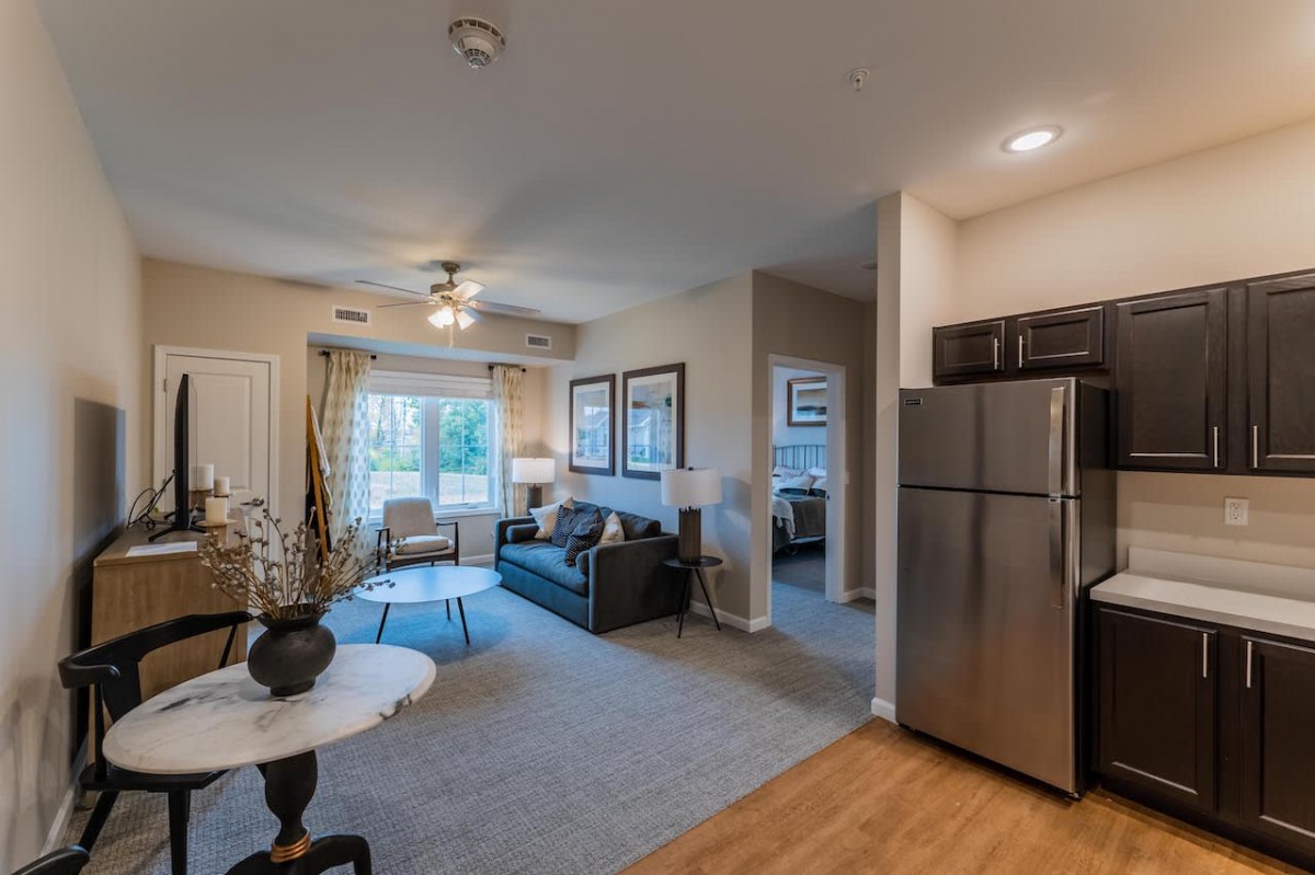A modern open floor plan apartment with a living room and kitchen area, featuring a comfortable sofa and stainless steel refrigerator.