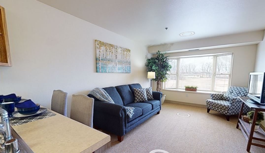 A cozy living room featuring a blue sofa, artwork on the wall, and natural light streaming through a large window.