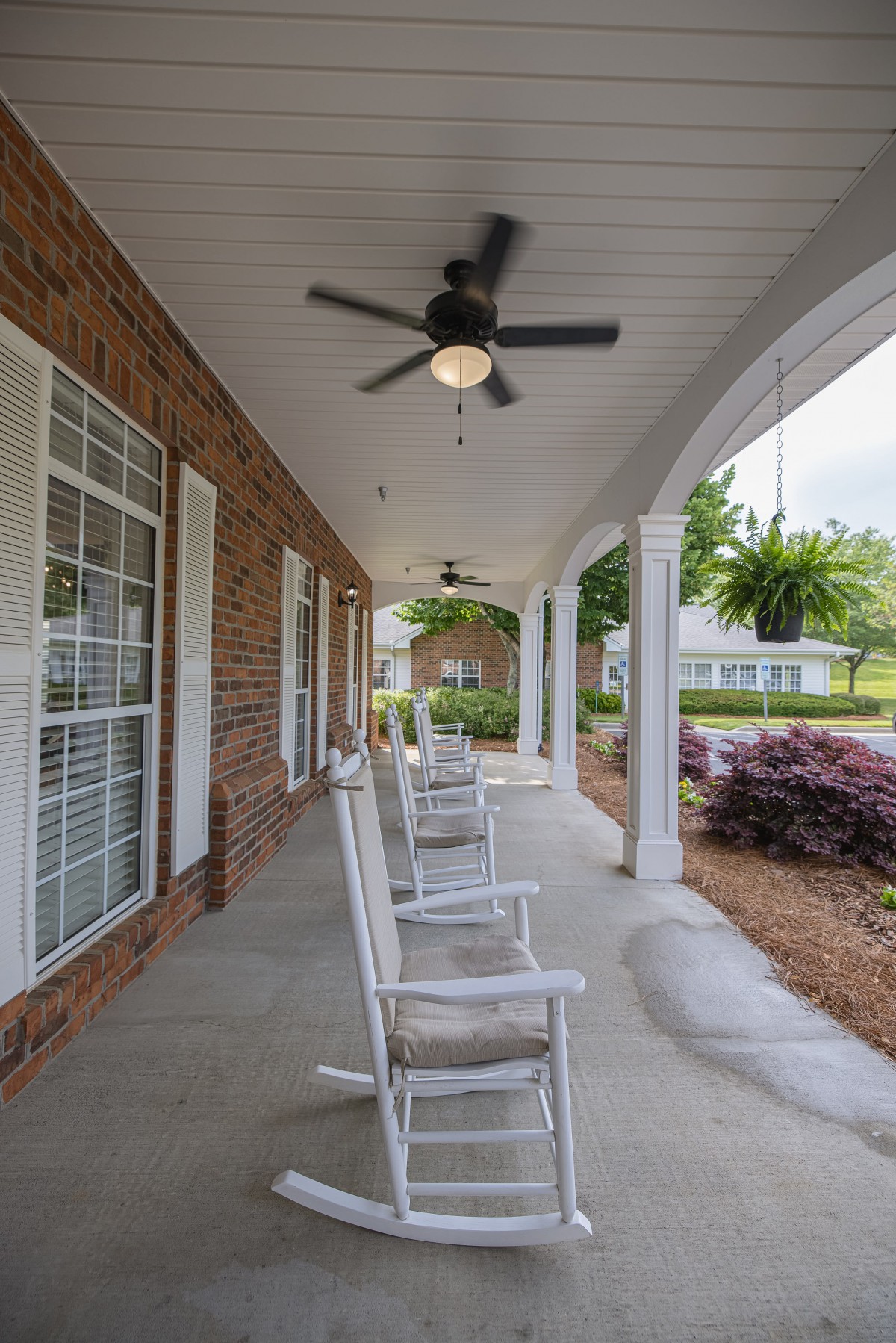A covered porch featuring rocking chairs, ceiling fans, and a brick exterior with lush landscaping.