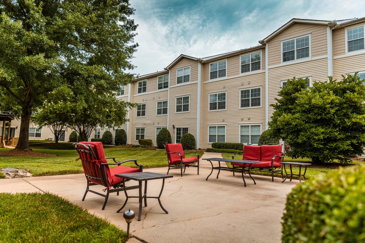 A well-maintained patio area with red chairs in front of a residential apartment building, surrounded by lush greenery.