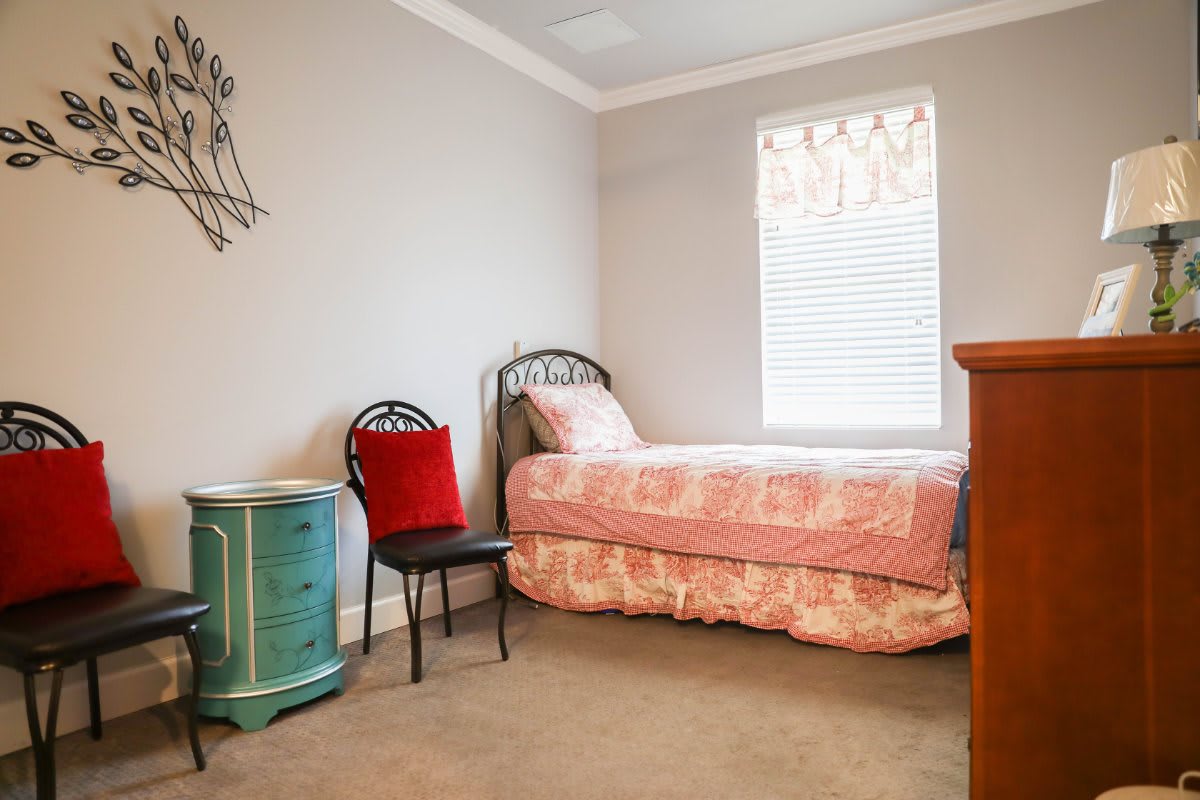 A cozy bedroom with a single bed, decorated in pink tones, featuring a window and an adjacent chair with a red pillow.