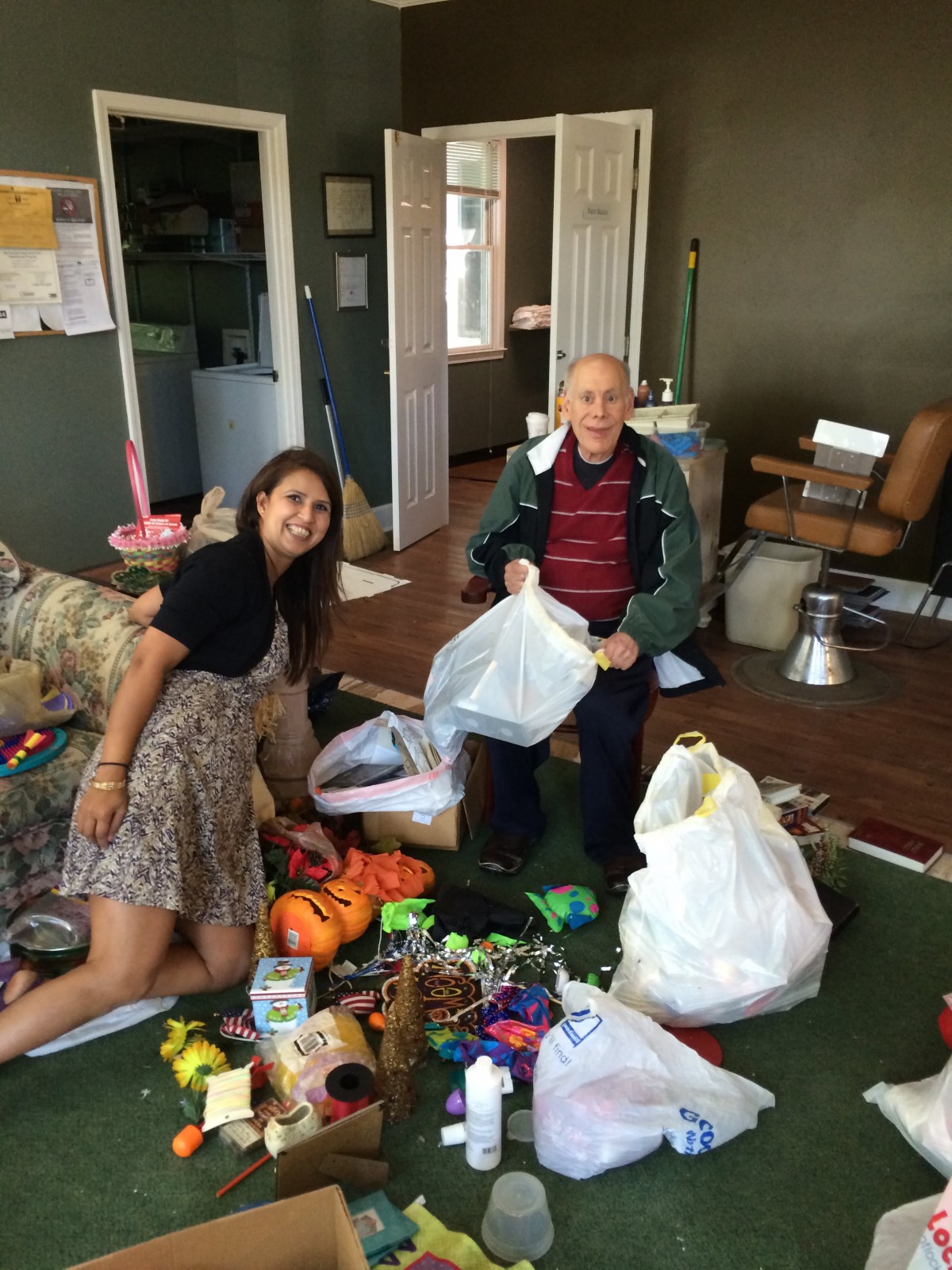 Two people organizing and cleaning a room filled with toys and bags.