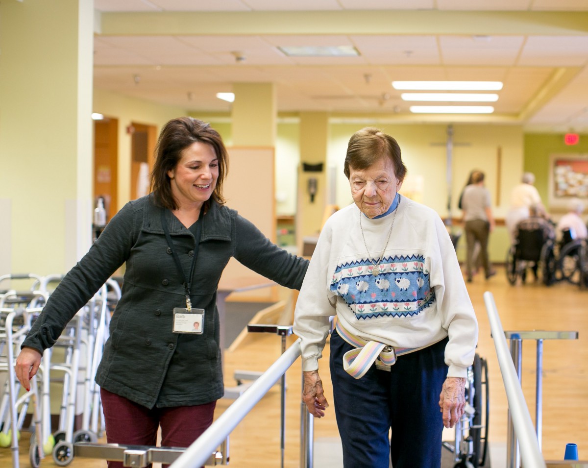 A healthcare professional assists an elderly woman with walking exercises in a rehabilitation setting.