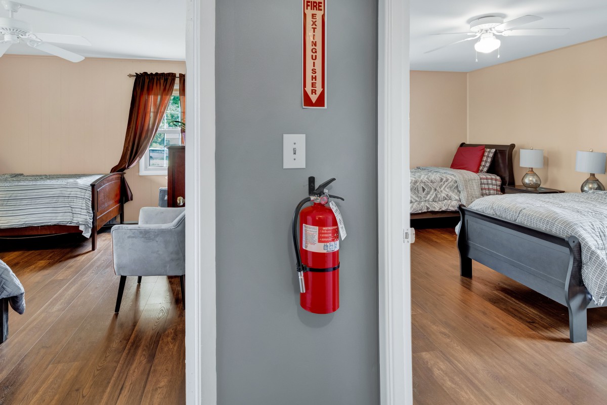 A hallway featuring a fire extinguisher mounted on the wall, leading to a bedroom with wooden flooring.