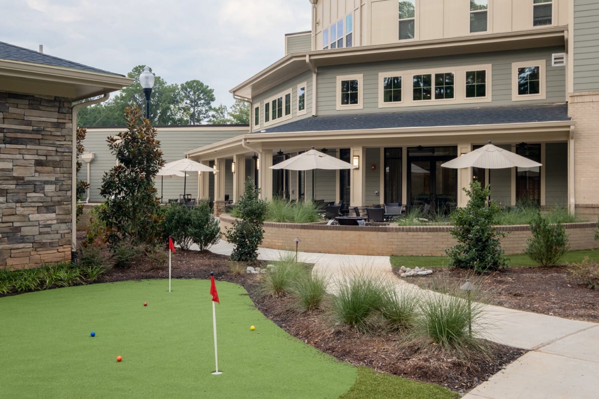 A view of a building with an outdoor patio featuring umbrellas and a golf putting green surrounded by landscaping.