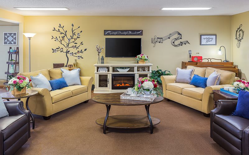 A cozy living room with two sofas, a coffee table, a TV above a fireplace, and wall art decorations.