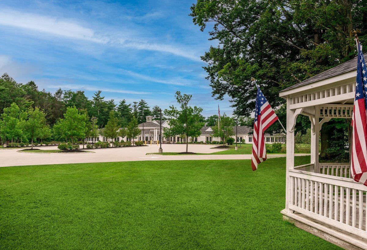 A picturesque park scene featuring a gazebo with an American flag, surrounded by lush green grass and trees under a clear blue sky.