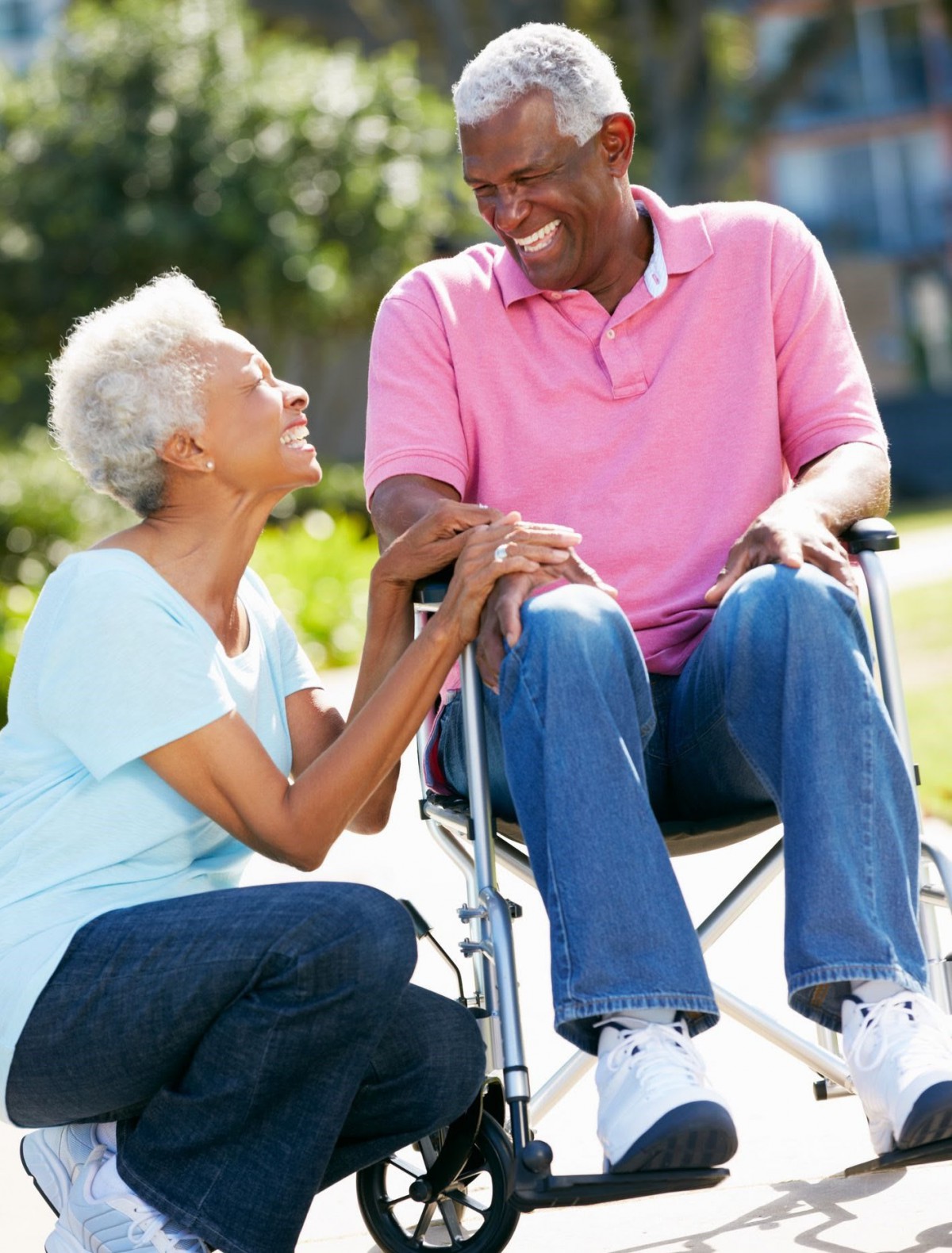 An elderly couple enjoys a sunny day outside, with one sitting in a wheelchair and both sharing a smile.