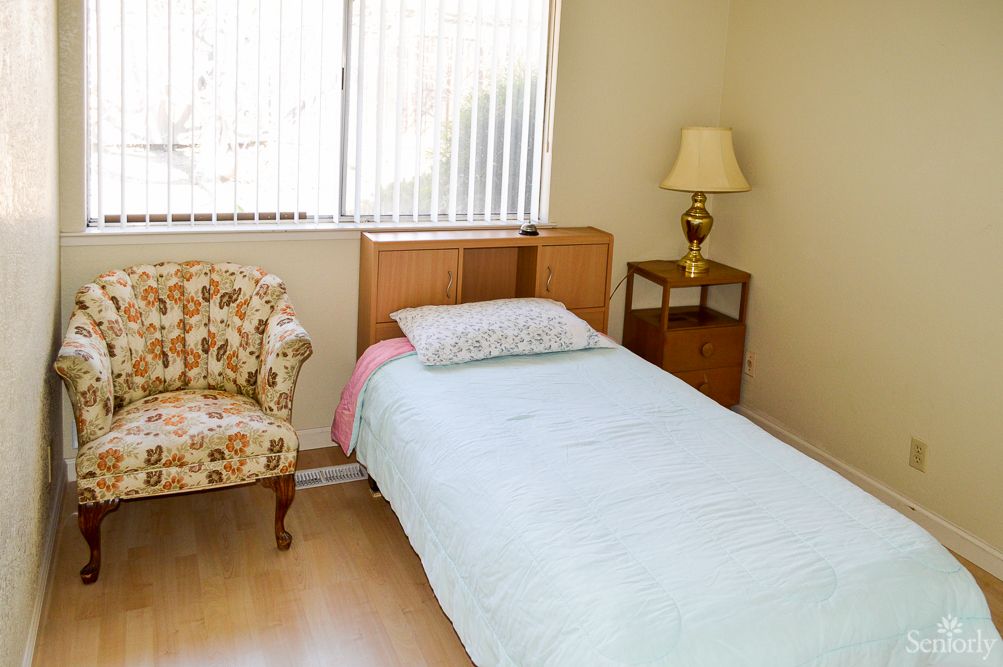 A simple bedroom with a single bed, floral chair, and a lamp on a nightstand by the window.