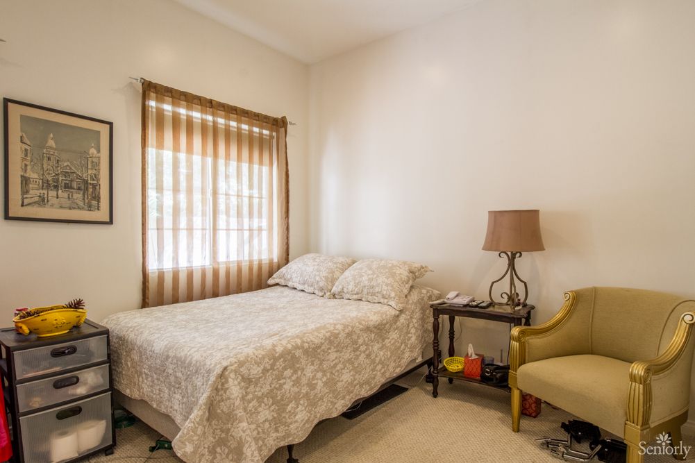 A cozy bedroom with a neatly made bed, a lamp on a nightstand, and a comfortable chair beside the window.