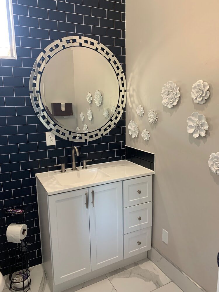A modern bathroom featuring a circular mirror with decorative edging, a white vanity with sink, and white floral wall decor against blue tiled walls.