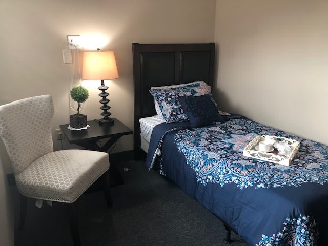 A cozy bedroom featuring a single bed with blue floral bedding, a small table with a lamp, and an upholstered chair.