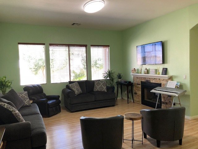 A cozy living room with green walls, featuring comfortable sofas, a television, and large windows letting in natural light.
