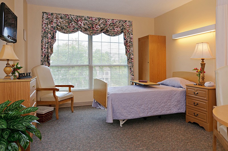 A hospital room with a single bed, chair, and large window with patterned curtains.