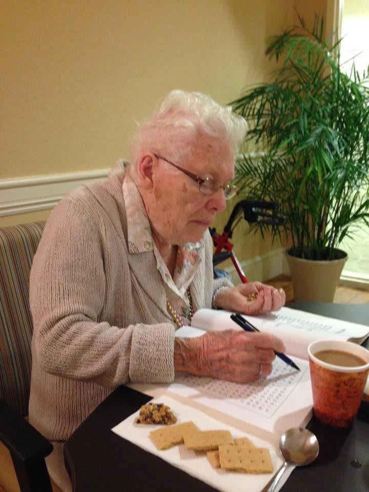 An elderly woman is seated at a table, focused on writing in a book with a pen. A cup and some snacks are on the table, and a plant is visible in the background.