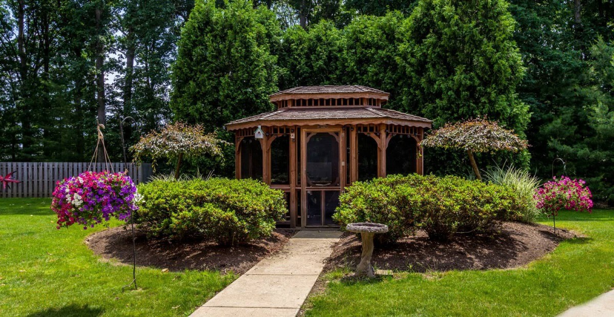 A wooden gazebo surrounded by lush greenery and bushes, with a pathway leading to its entrance.
