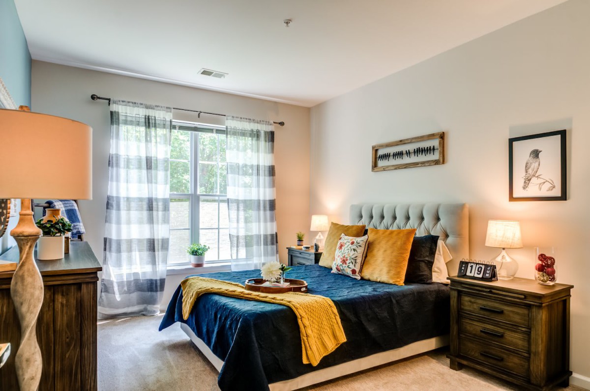 A cozy and modern bedroom with natural light filtering through checkered curtains, featuring a neatly arranged bed with blue and yellow accents.