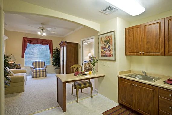 A cozy apartment interior featuring a small kitchen with wood cabinets and an adjoining dining area, opening into a living room.