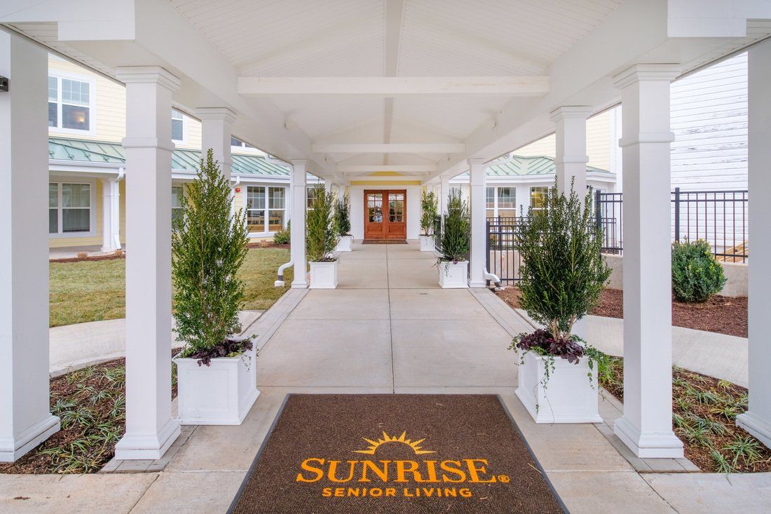 A covered walkway with potted plants leads to a building entrance, featuring a mat with the 'Sunrise Senior Living' logo.