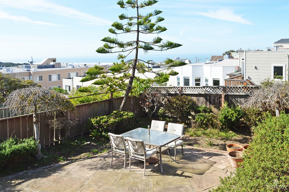 A cozy backyard patio with a glass table and chairs, surrounded by greenery and urban houses.