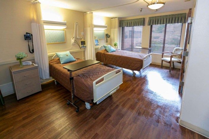 A clean and well-lit hospital room featuring two patient beds with large windows providing natural light.