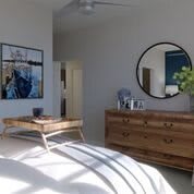 A modern bedroom featuring a wooden dresser with a round mirror, a neatly made bed with a tray on top, and artwork on the wall.