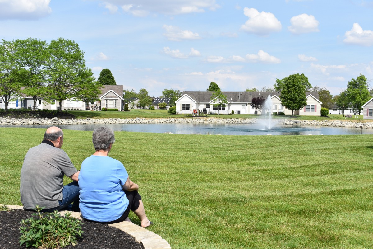 Two people sitting near a pond with a fountain, surrounded by grass and houses.