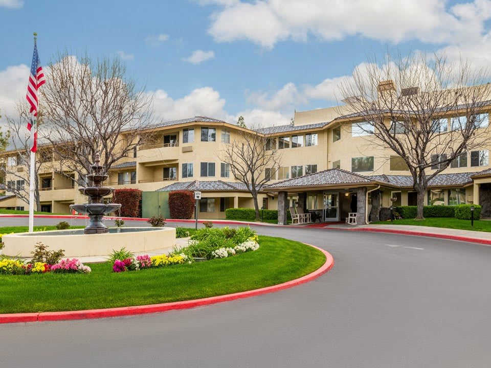 A beautifully landscaped residential building with a driveway and fountain on a cloudy day.