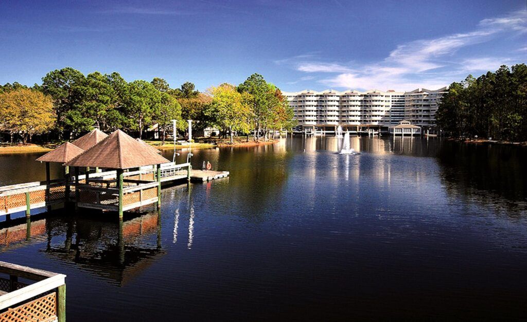 A serene lakeside view with a boat dock, fountain, and modern buildings in the background.