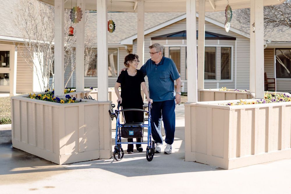 A caregiver assists an elderly person using a walker outside a building.