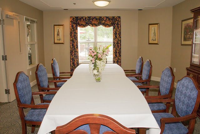 A neatly arranged dining room with a long table and chairs, featuring decorative curtains and a flower centerpiece.