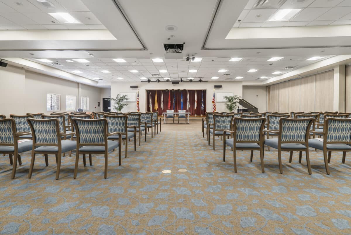 An empty conference room with arranged chairs facing a podium and flags.
