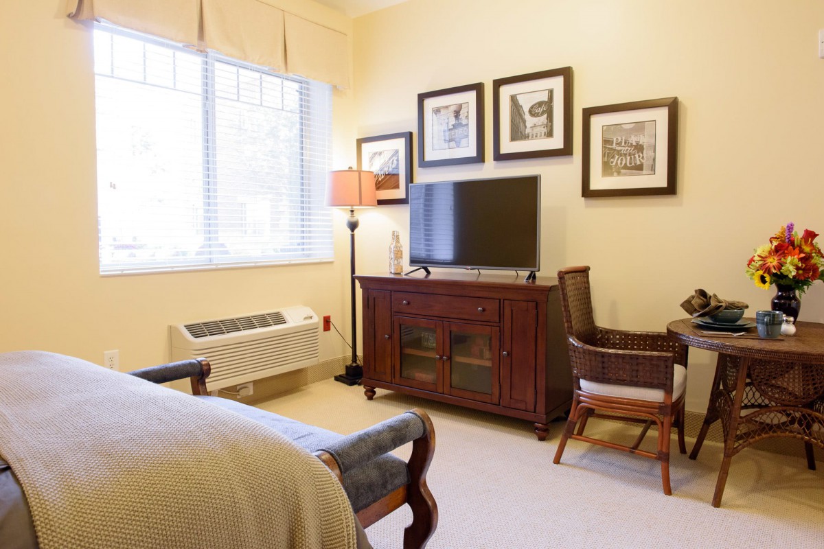 A cozy bedroom featuring a television on a wooden cabinet, with artwork on the walls and natural light streaming through the window.