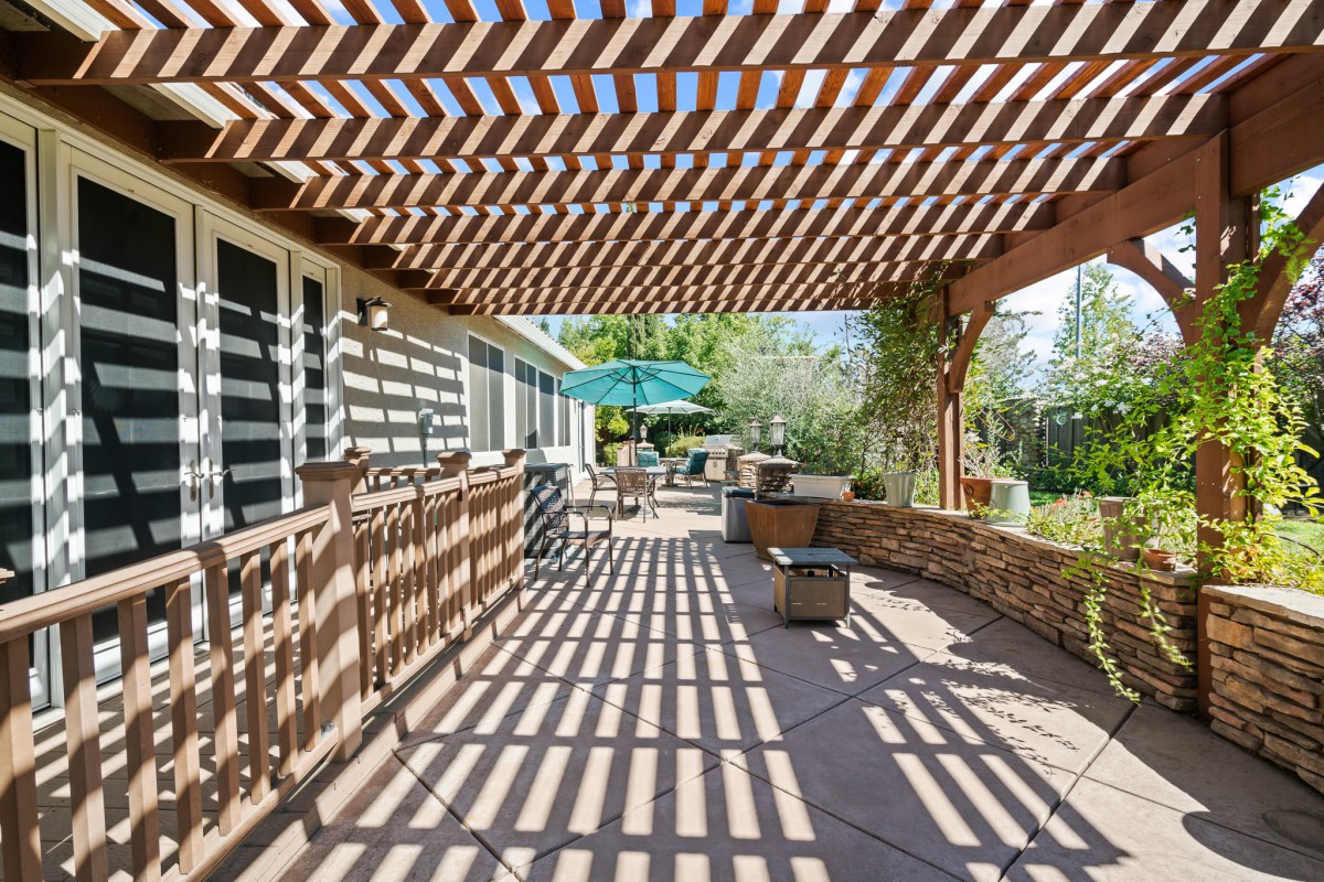 A sunny patio with a wooden pergola casting shadows, featuring outdoor furniture and garden views.