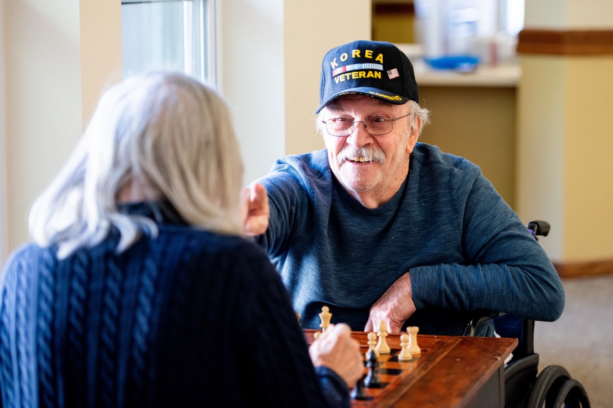 Two elderly individuals playing chess indoors, one wearing a veteran cap.